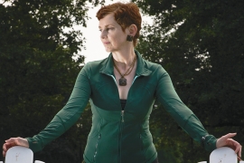 Catherine Waters Raymond, a woman with red hair wearing a green shirt, stands between two white chairs.